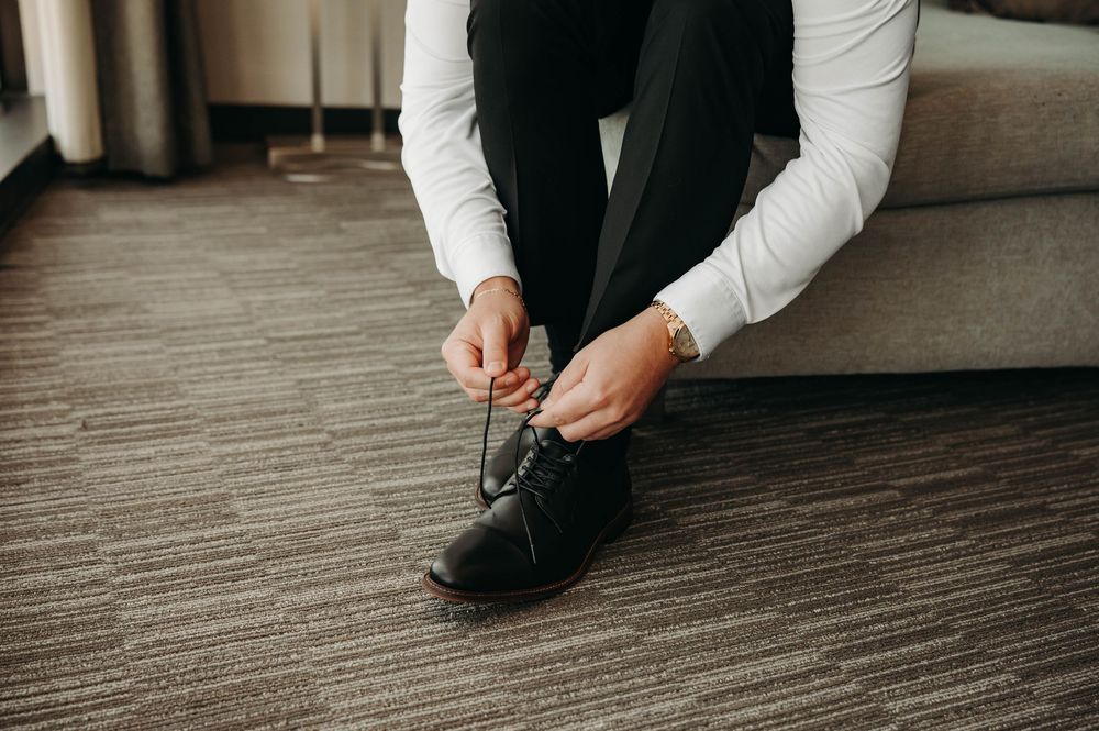 Groom tying black oxford dress shoes while seated, wearing black trousers and white dress shirt with gold watch.
