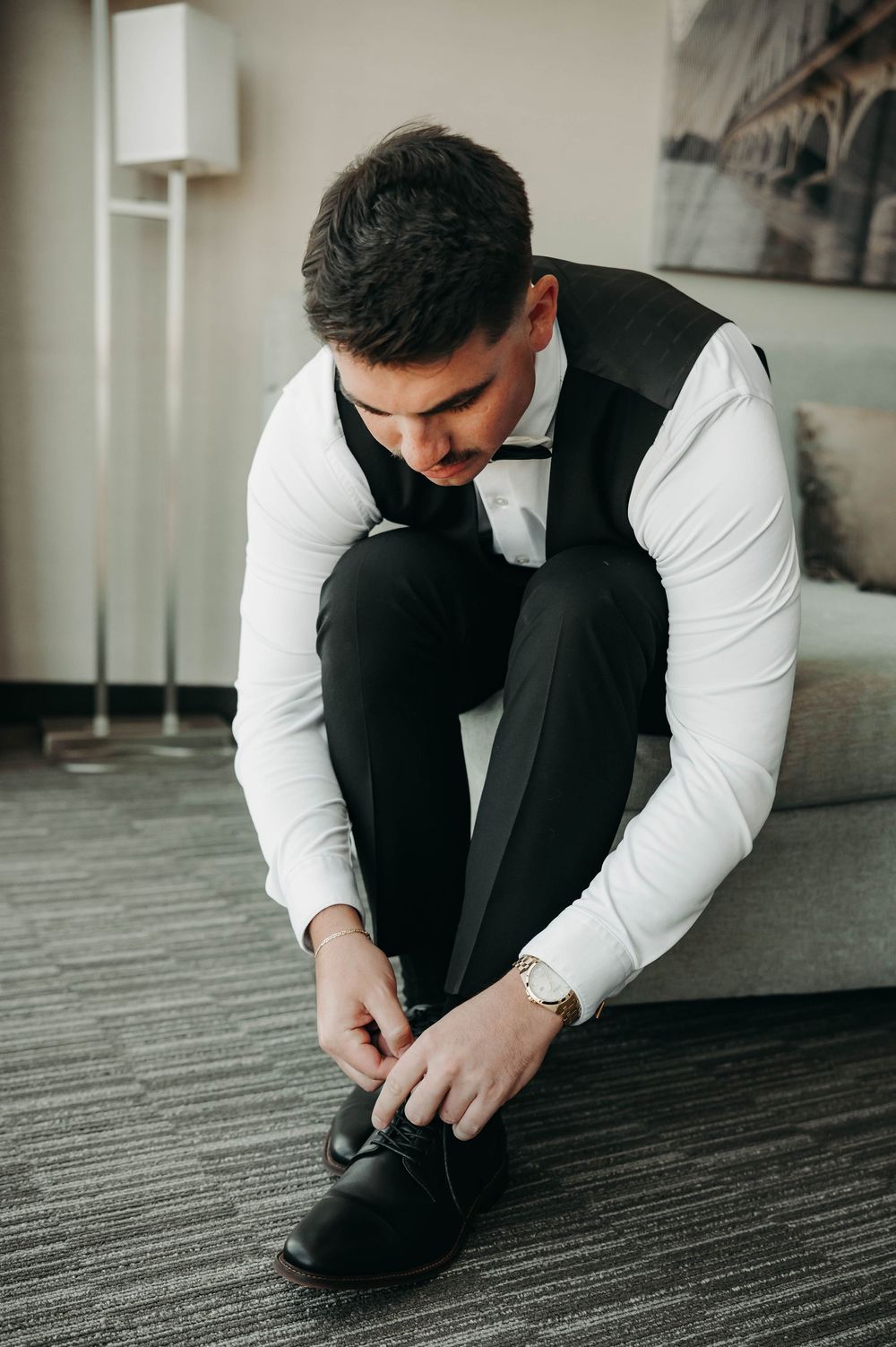 Dapper groom in black tuxedo vest and bow tie tying his dress shoes while seated on hotel room couch.