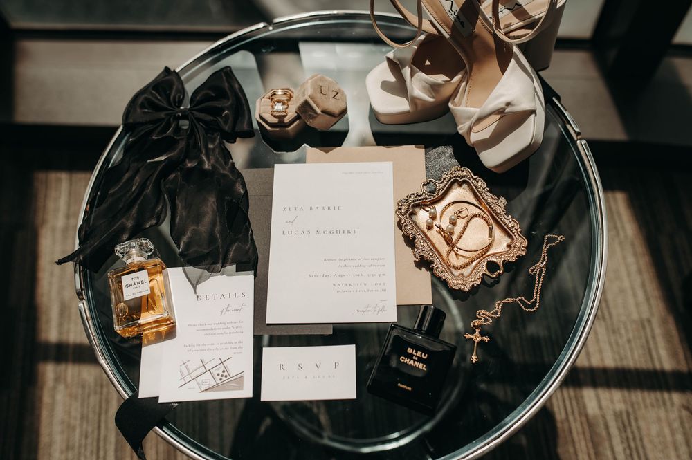 Elegant wedding flat lay featuring invitations, perfume, heels, jewelry, and black Chanel accessories.