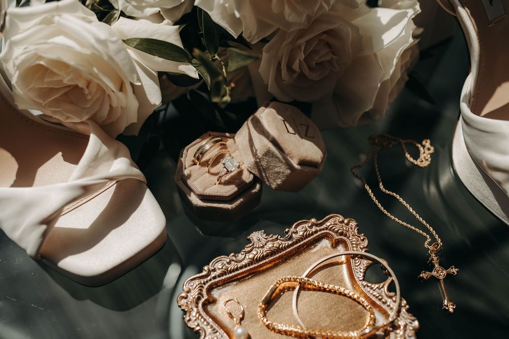 Bridal flat lay with white roses, gold jewelry, cross necklace, bracelets, and strappy heels.