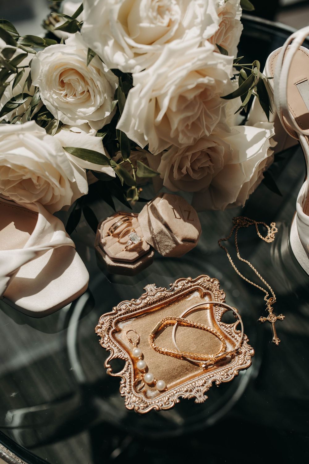 Elegant bridal flat lay featuring white roses, gold cross necklace, pearl bracelets, and strappy heels.