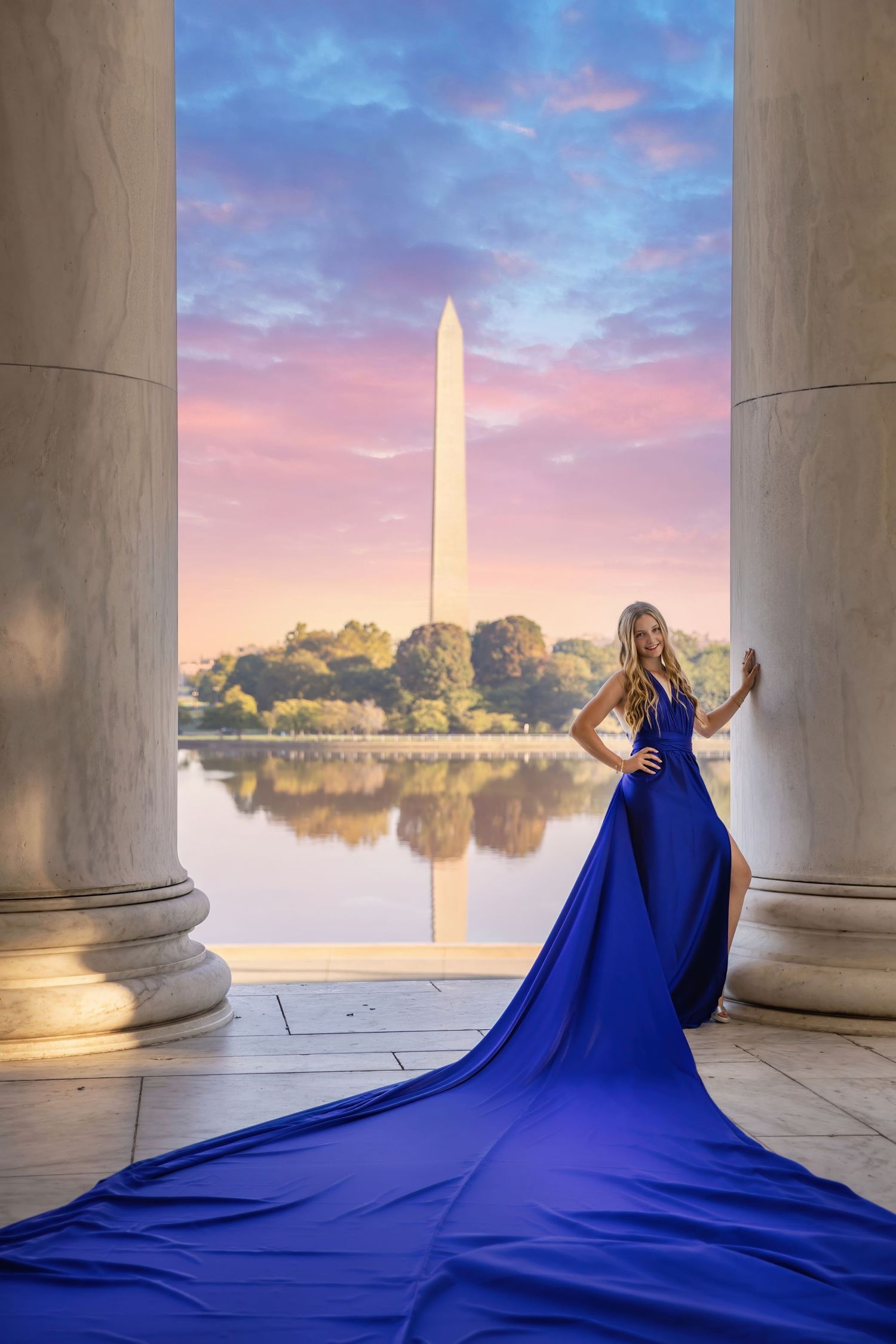 A flowing royal blue dress train spreads dramatically between marble columns with the Washington Monument at sunrise.
