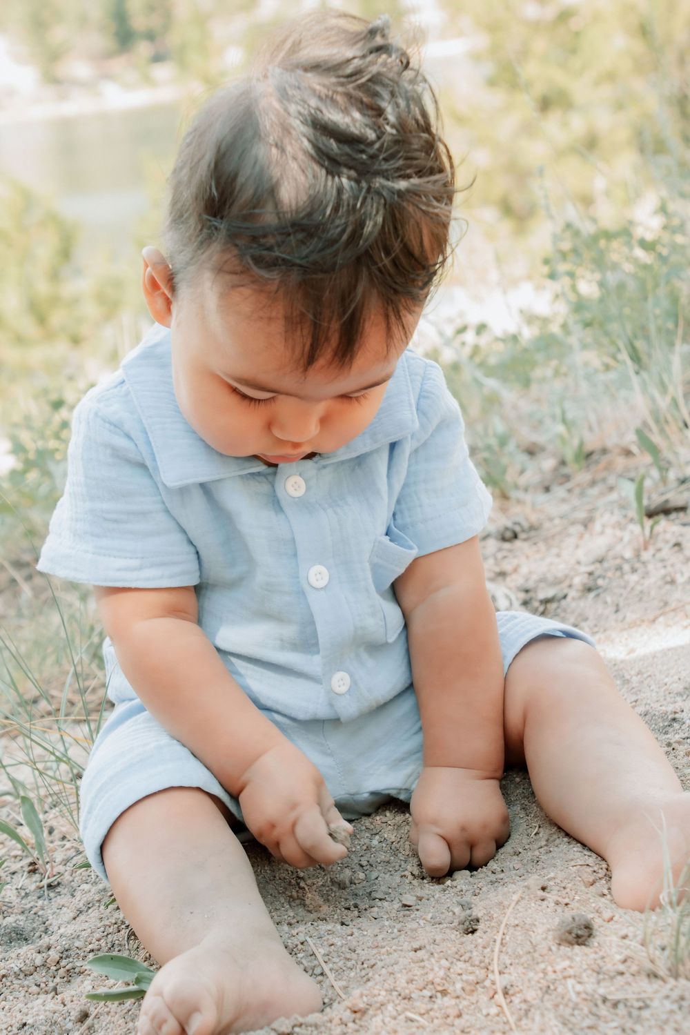 a baby sitting near lake looking down at hands playing with dirt