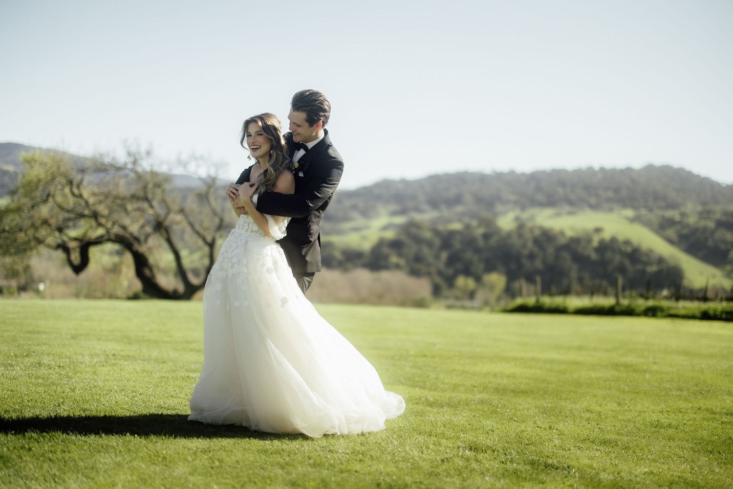 A couple in formal wedding attire stands together on a vast green golf course at sunset.