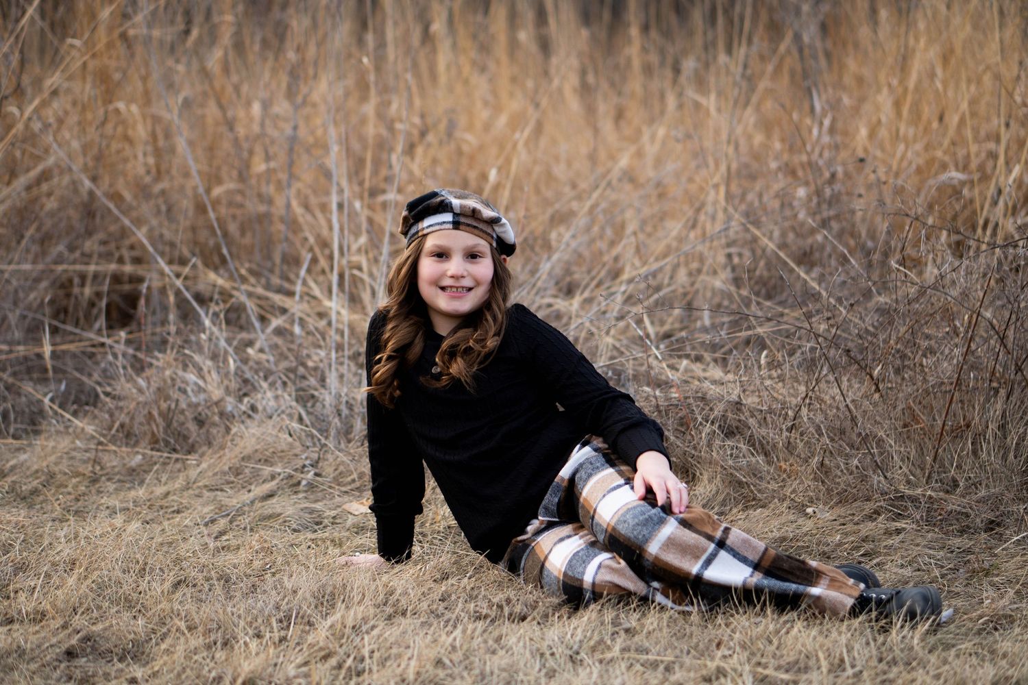 A young girl in a plaid skirt and beret sits smiling in a golden field of tall dry grass.