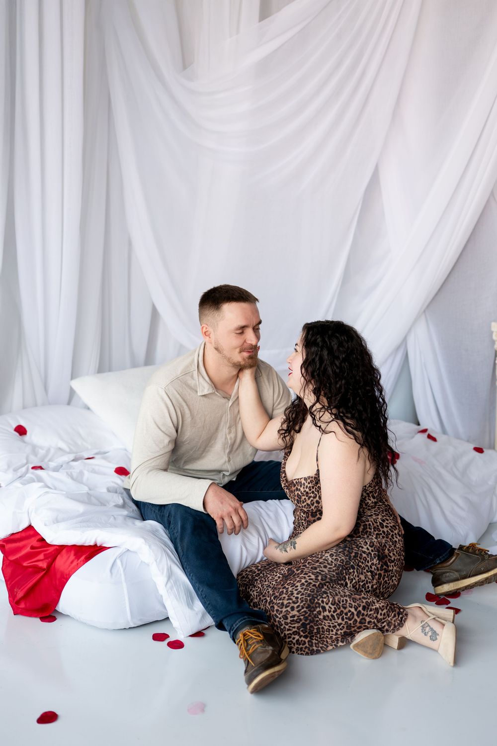 Loving couple sitting on the floor by a canopy bed with rose petals, gazing into each other's eyes.