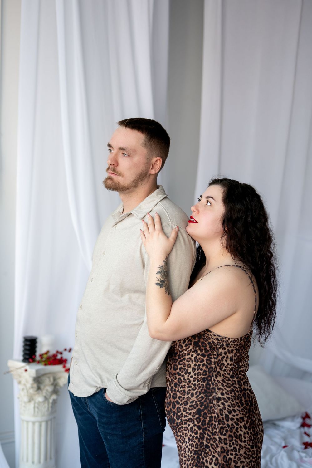Couple posing together, woman in leopard print dress touching man's shoulder as he looks away in white studio.