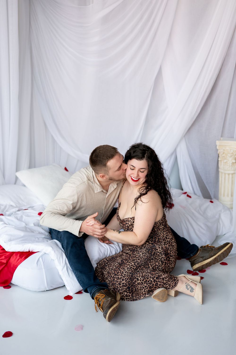 A man kisses his smiling partner on the cheek as they sit together on a bed surrounded by rose petals.