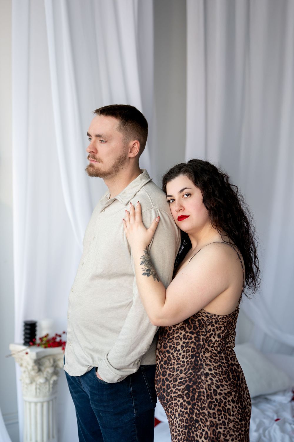 Couple standing back to back, woman in leopard print dress and man in beige shirt posing in white studio.
