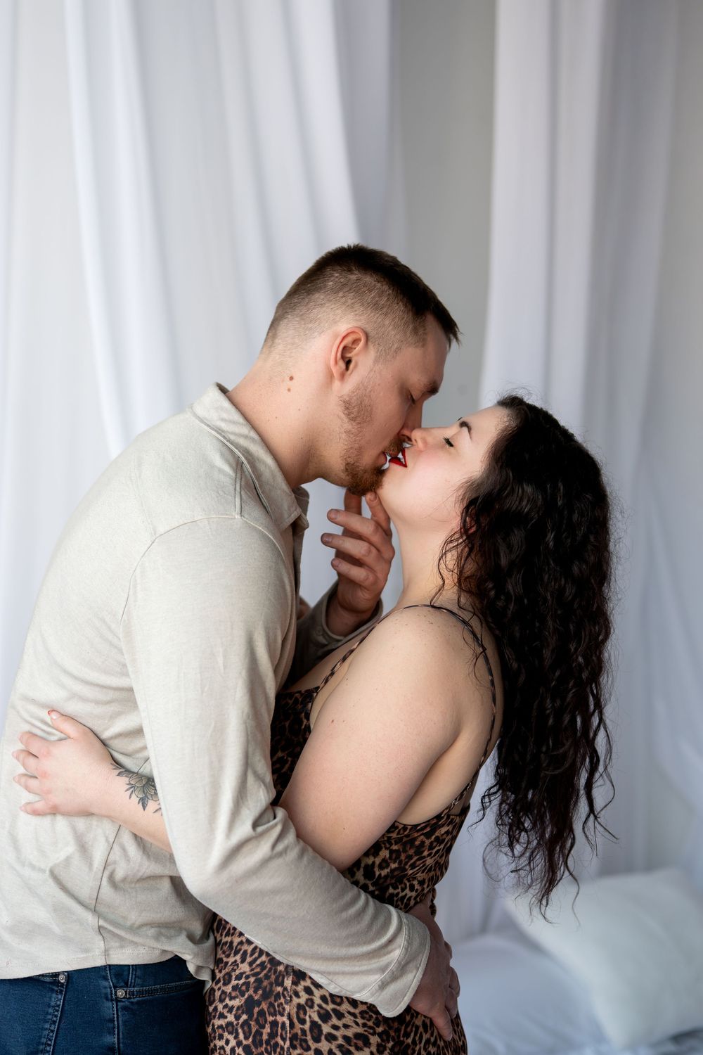 Couple kissing and embracing, woman in leopard print dress with red lips and man in beige shirt in white studio.