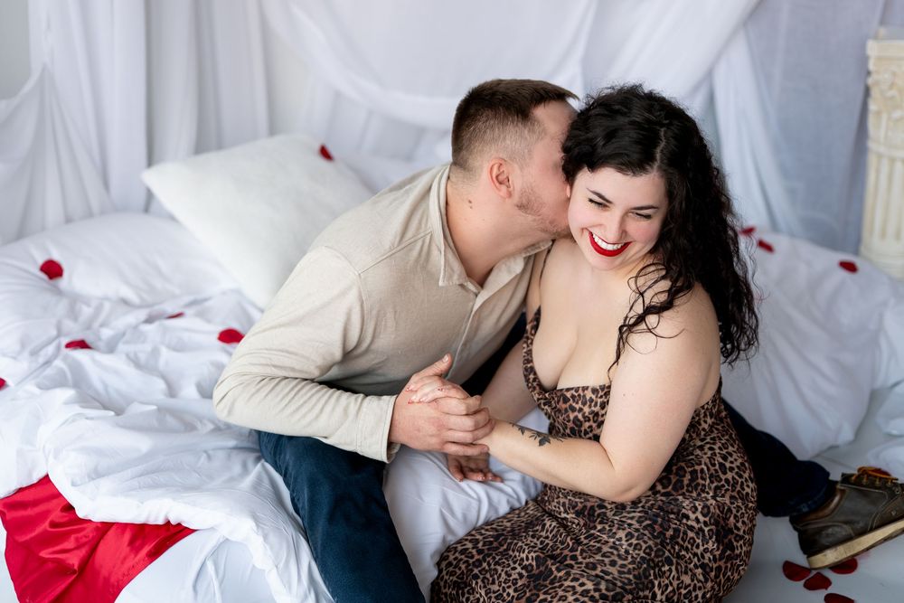 Man kissing his laughing partner on the cheek while sitting on a bed decorated with red rose petals.