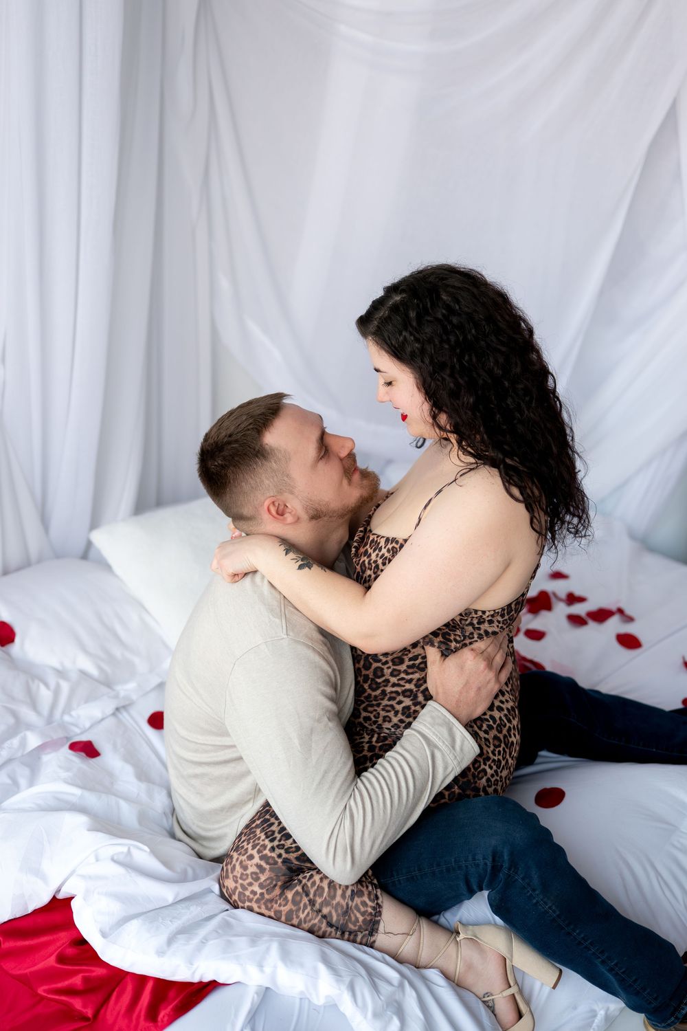 Romantic couple embracing on a white bed decorated with rose petals in a bright studio setting.