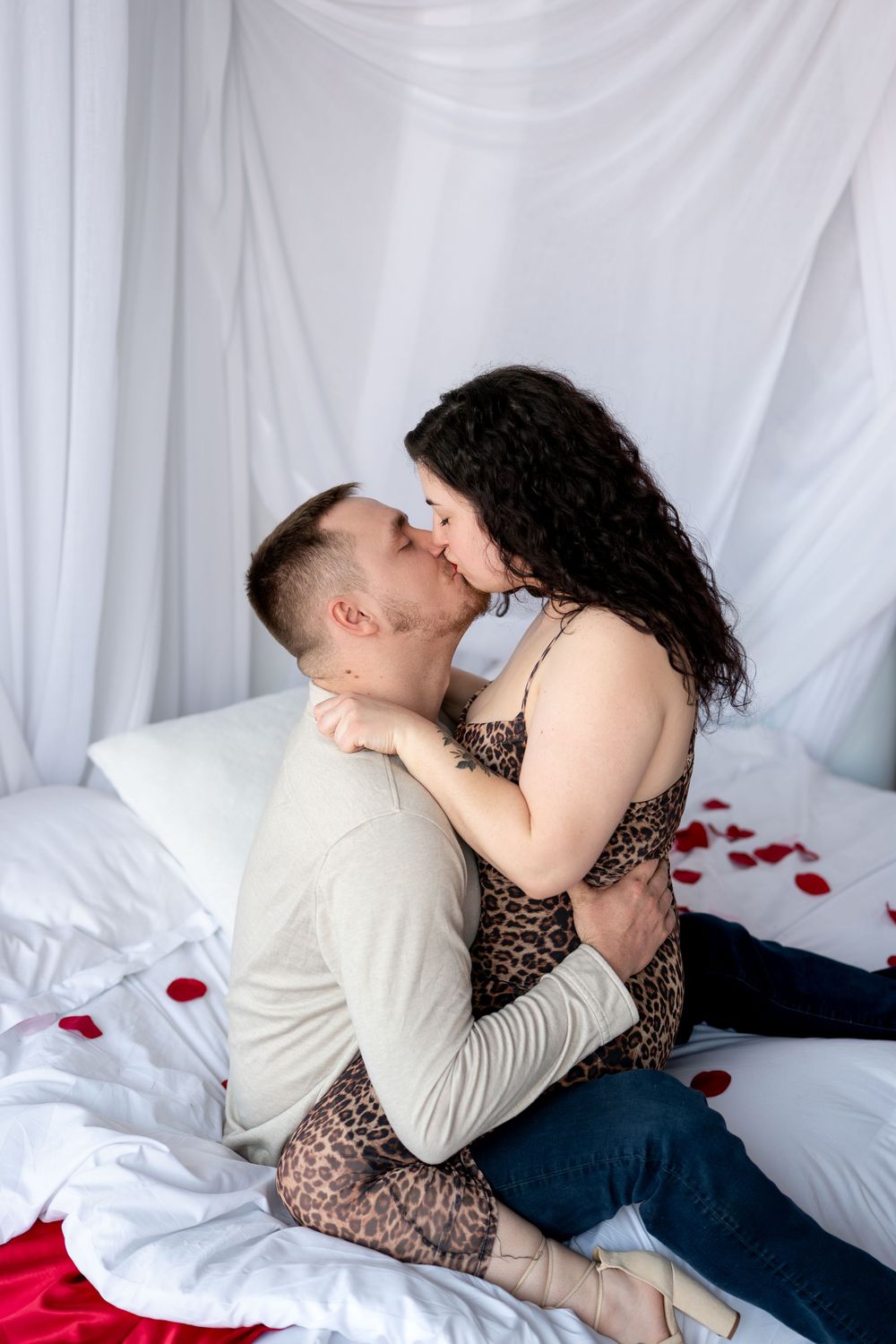 Passionate couple sharing a kiss while sitting on a white bed adorned with scattered red rose petals.