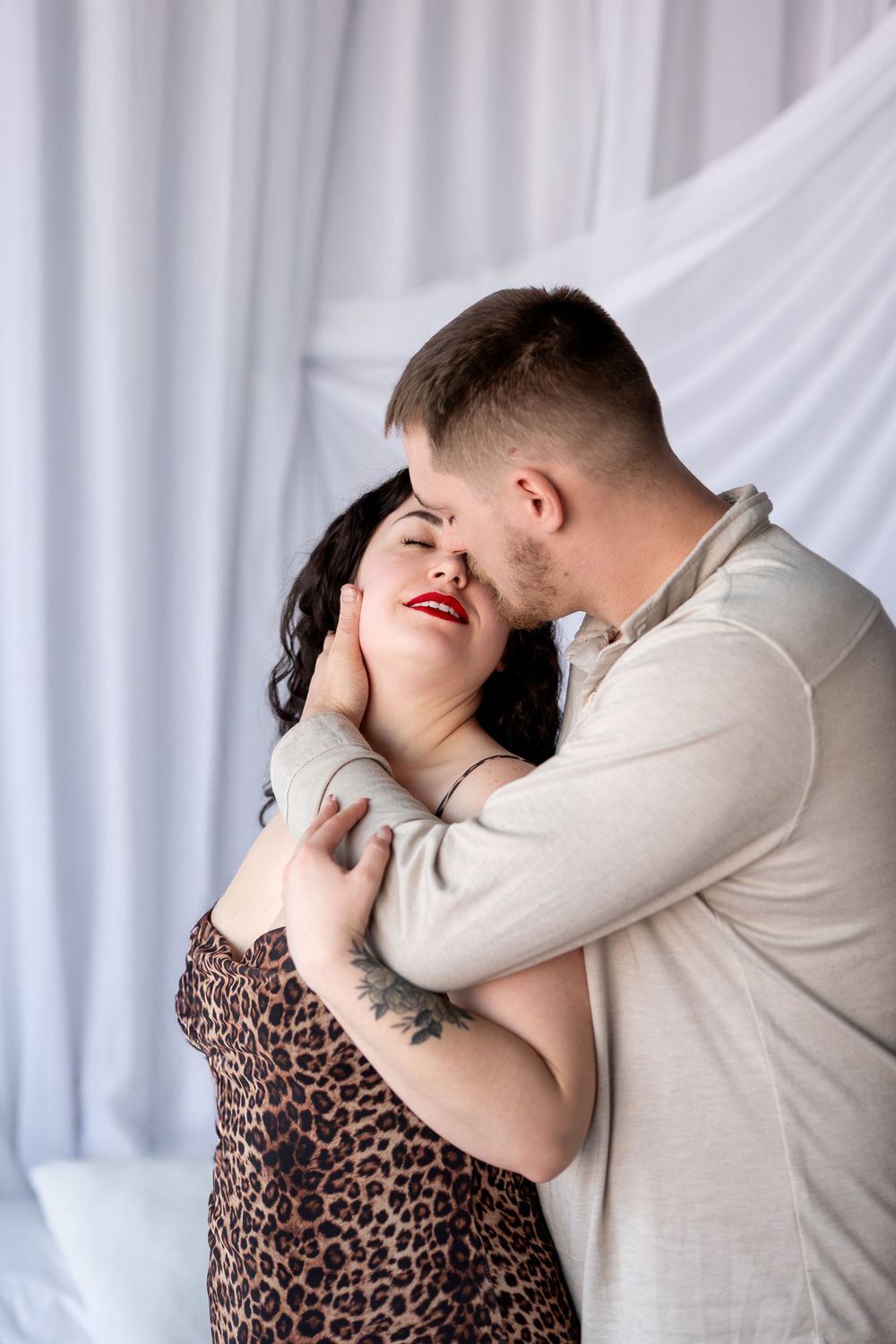 Man tenderly kisses woman in leopard dress while embracing her from behind against white curtain backdrop.