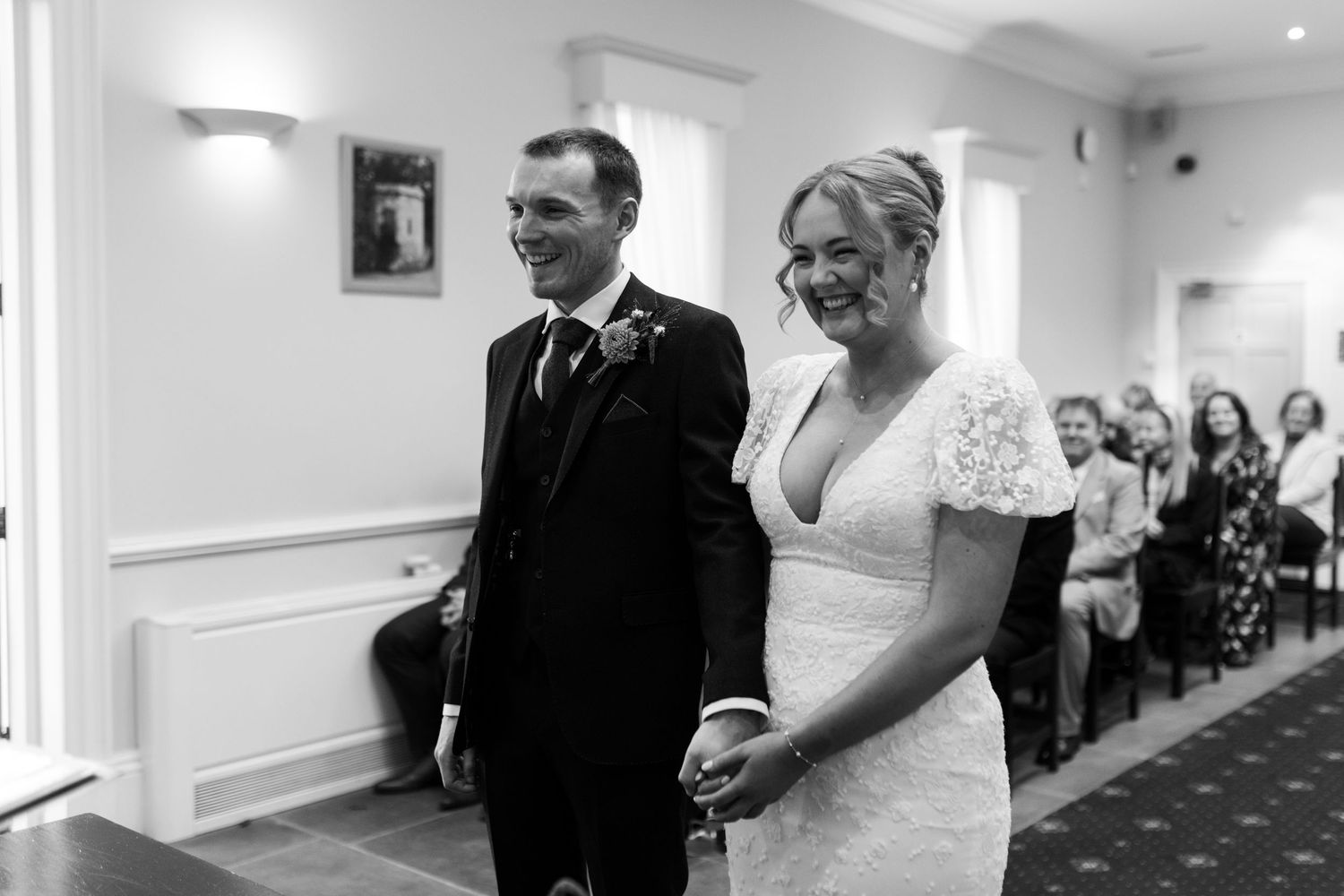 A black and white wedding ceremony photo showing a couple standing together at the altar in a bright indoor venue.