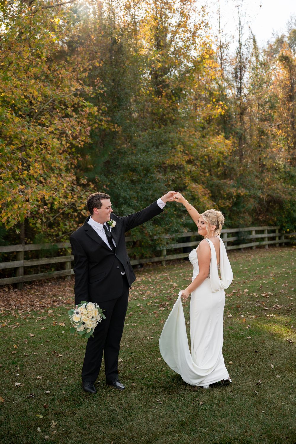 Wedding couple dancing playfully on lawn with autumn foliage and rustic fence in background.