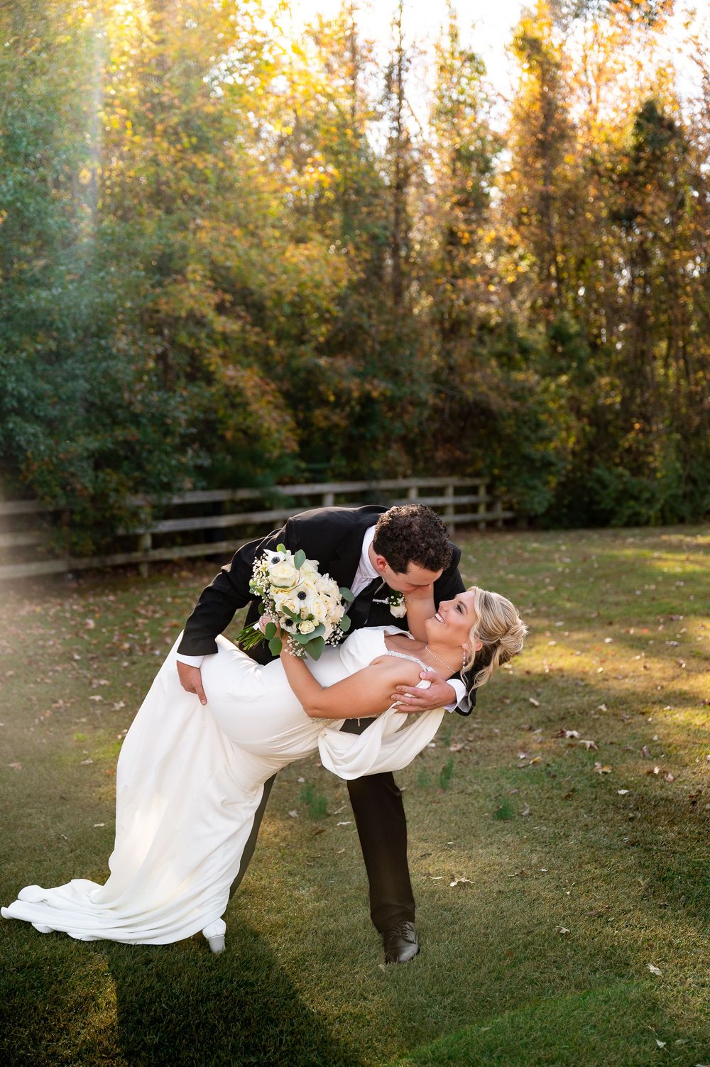 Dramatic wedding dip pose with autumn trees and wooden fence in background.