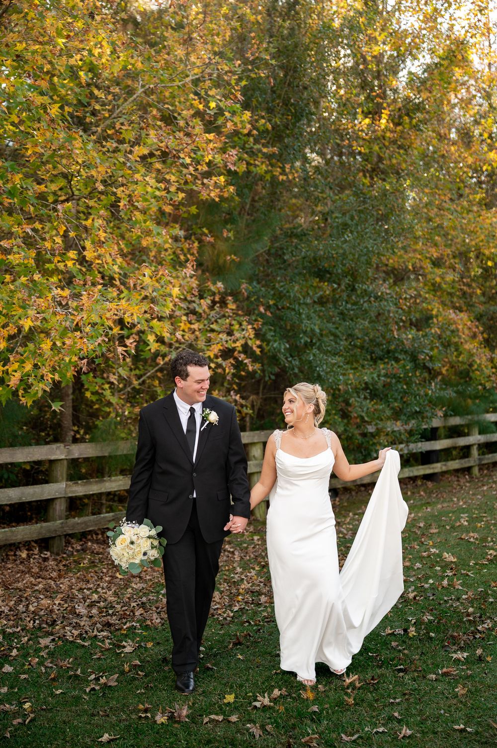 Elegant couple walking together on lawn with fall trees and white bouquet.