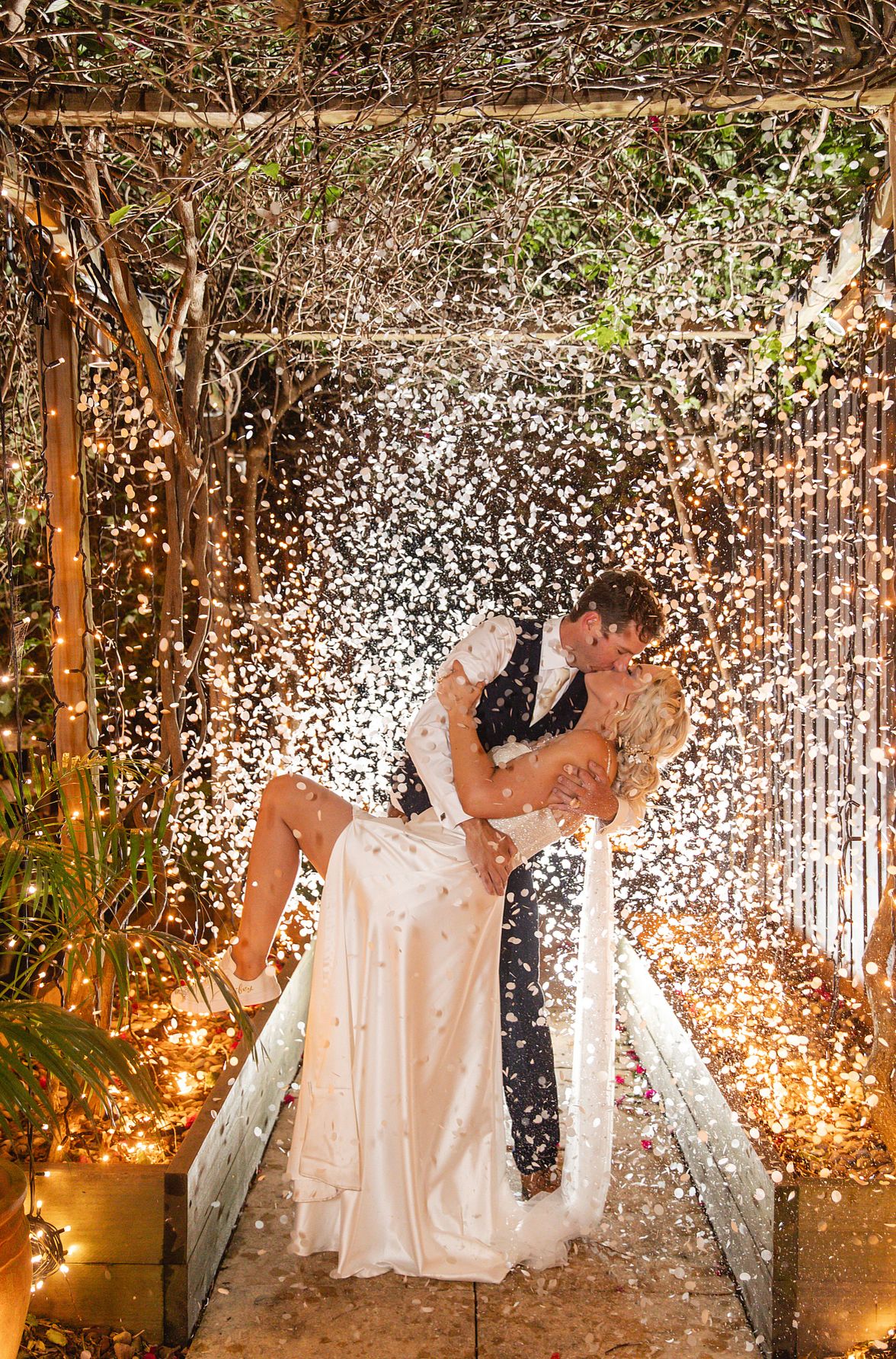 Groom lifts bride in a passionate kiss under a shower of confetti and fairy lights at night.