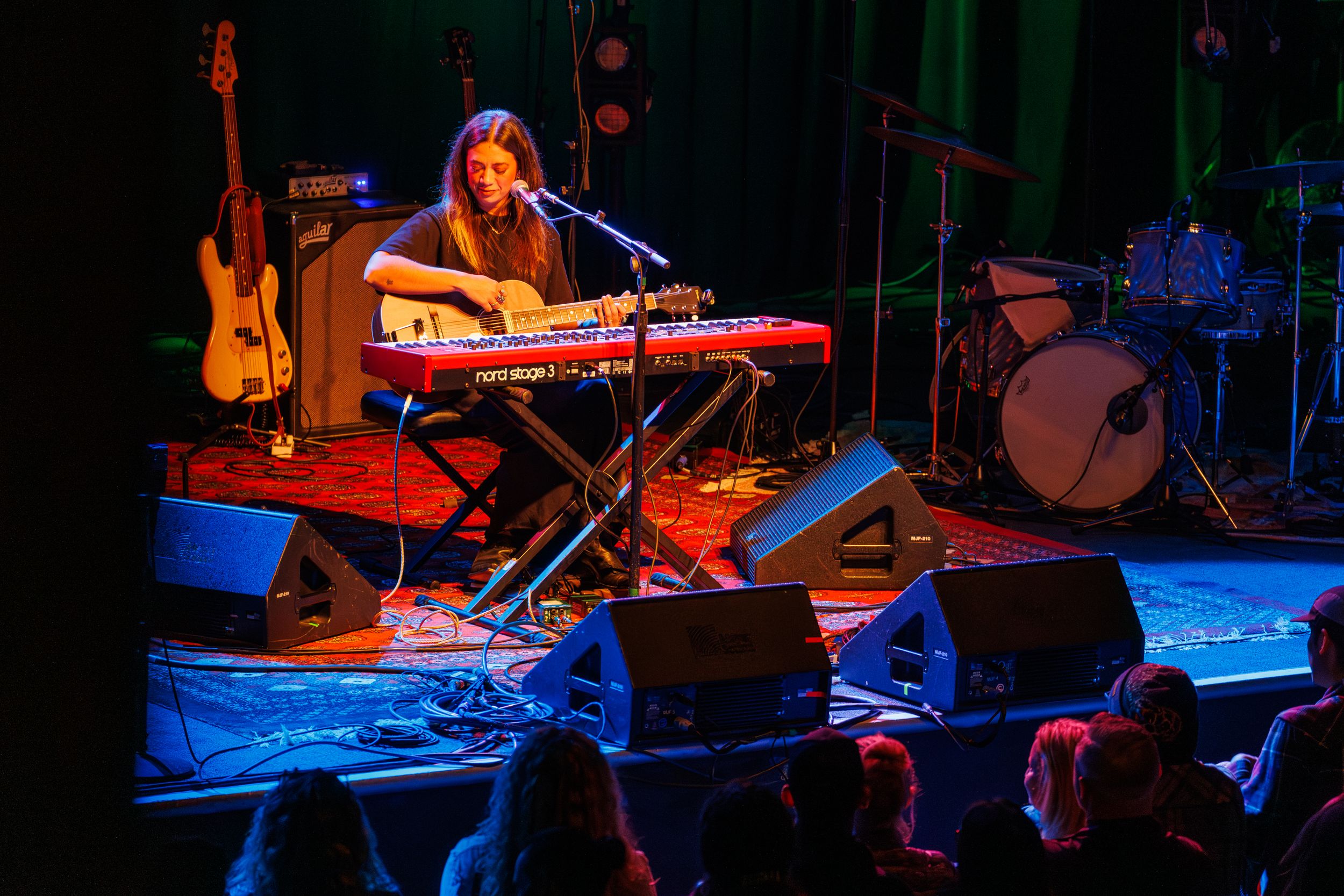 Musicians perform on a dimly lit stage with blue lighting and various instruments including keyboards and guitars.
