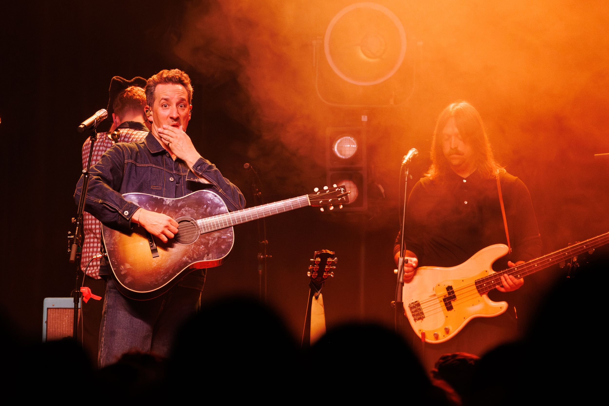 Musicians perform on stage with guitars under dramatic orange and red lighting during a live concert performance.