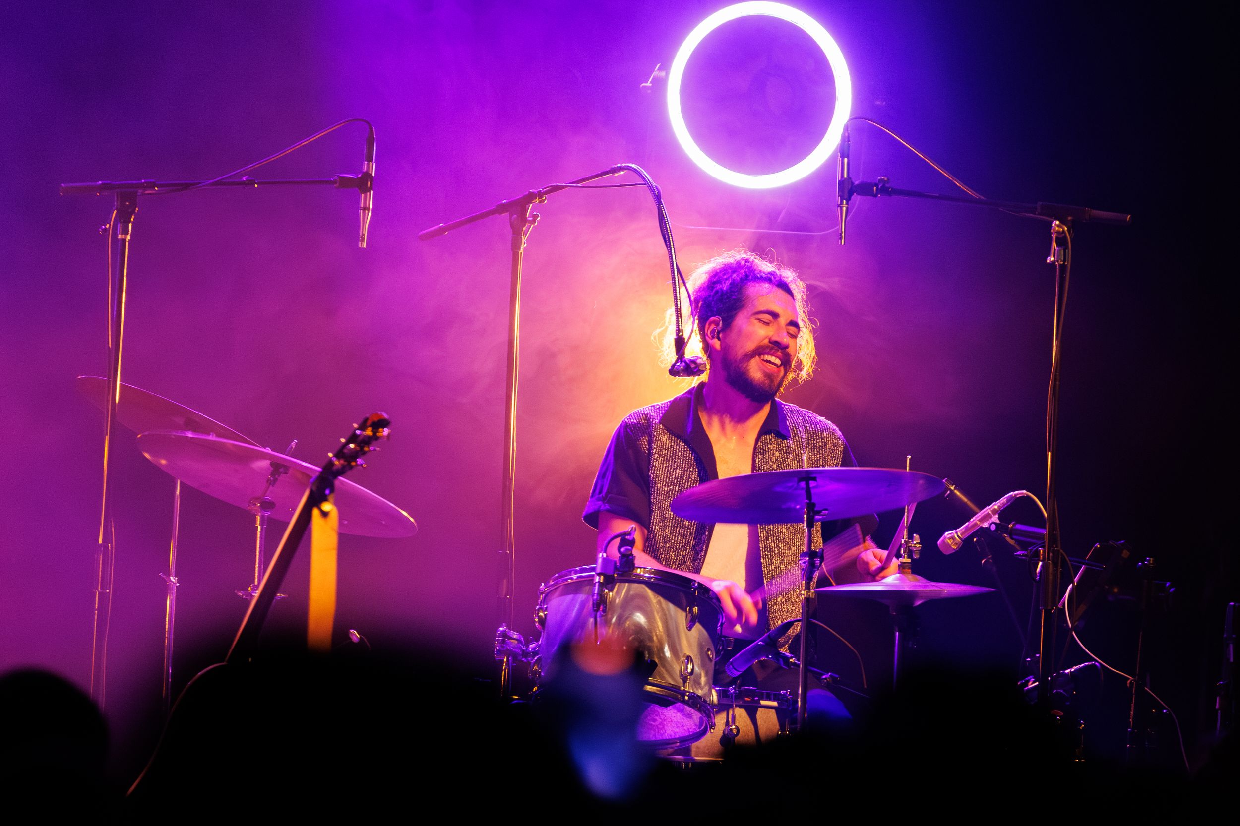 Musicians perform on stage with vibrant purple and pink lighting creating a dramatic concert atmosphere.
