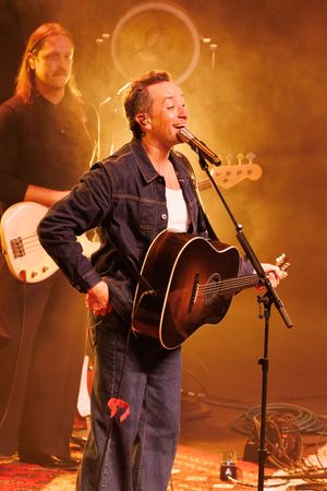 Musician performs on stage with acoustic guitar wearing denim jacket in warm golden lighting with band member in background.