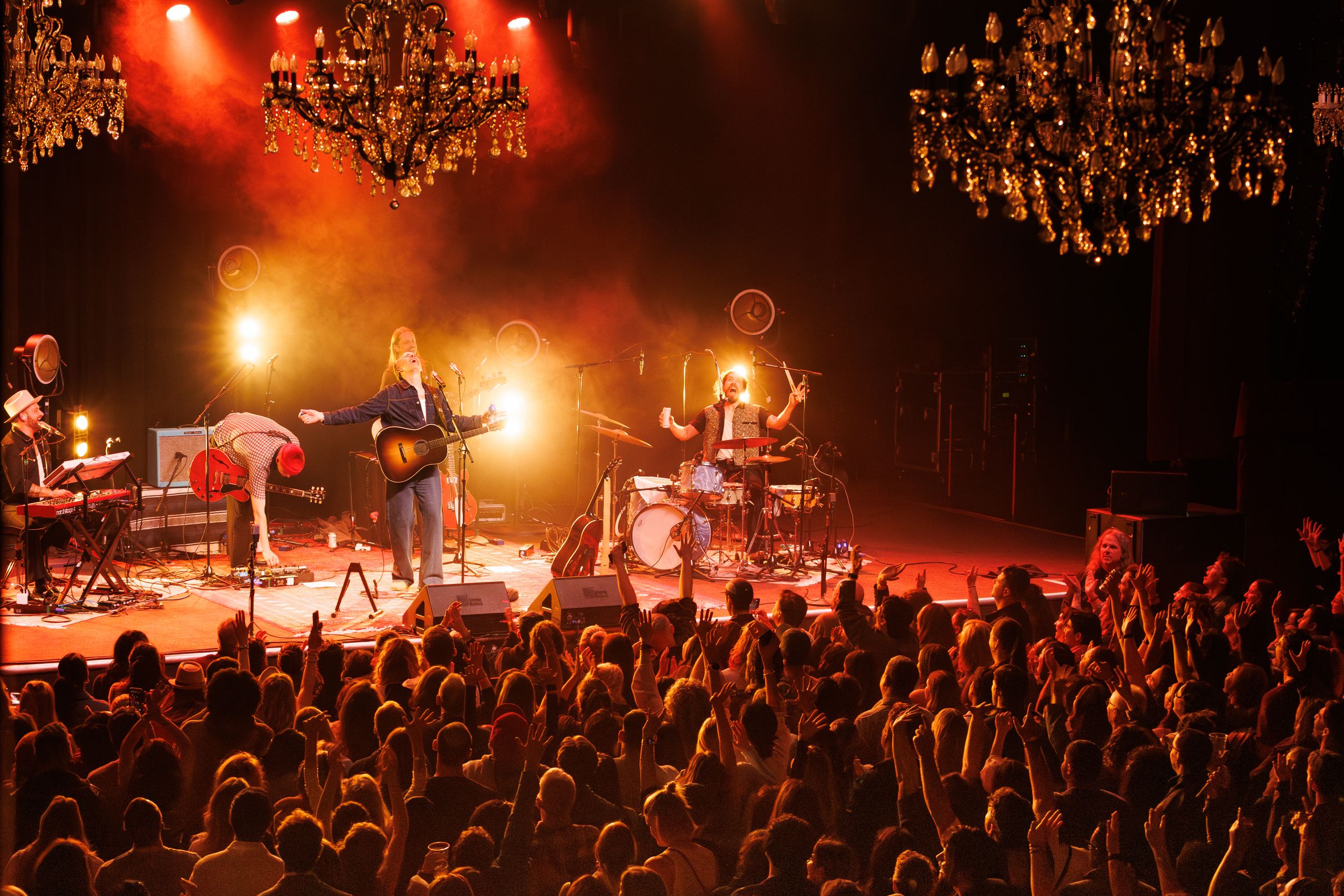 Rock band performs on stage under red lighting and chandeliers as crowd cheers at concert venue.