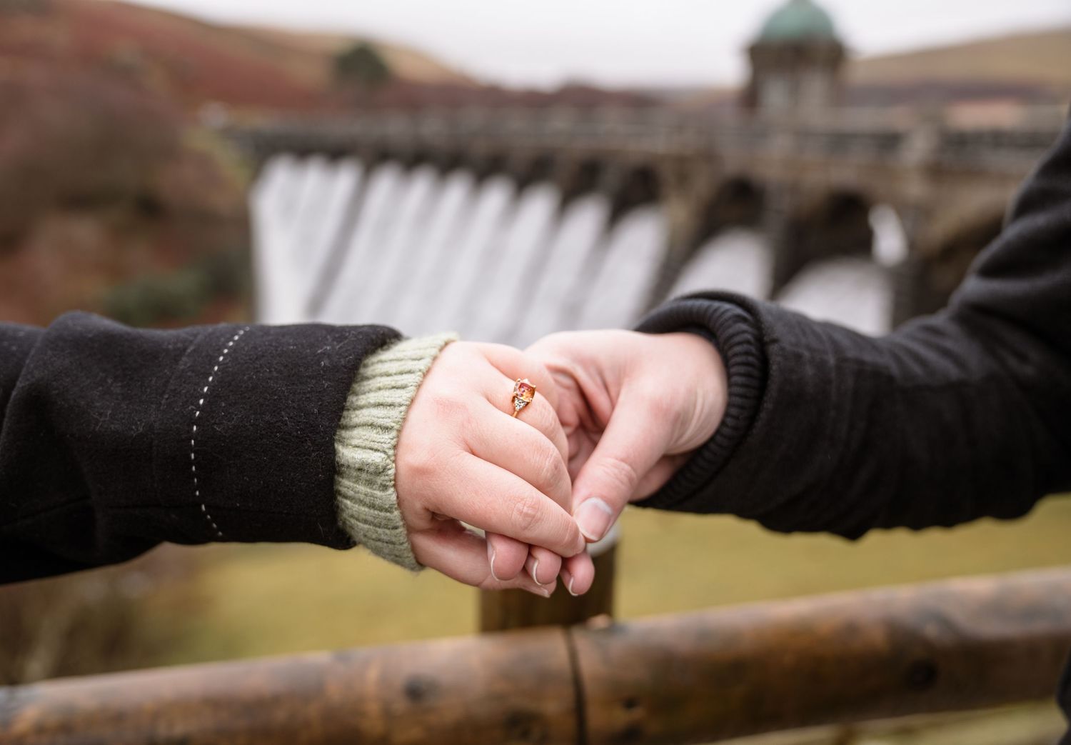 Two people hold hands while leaning on a wooden railing in an outdoor setting.