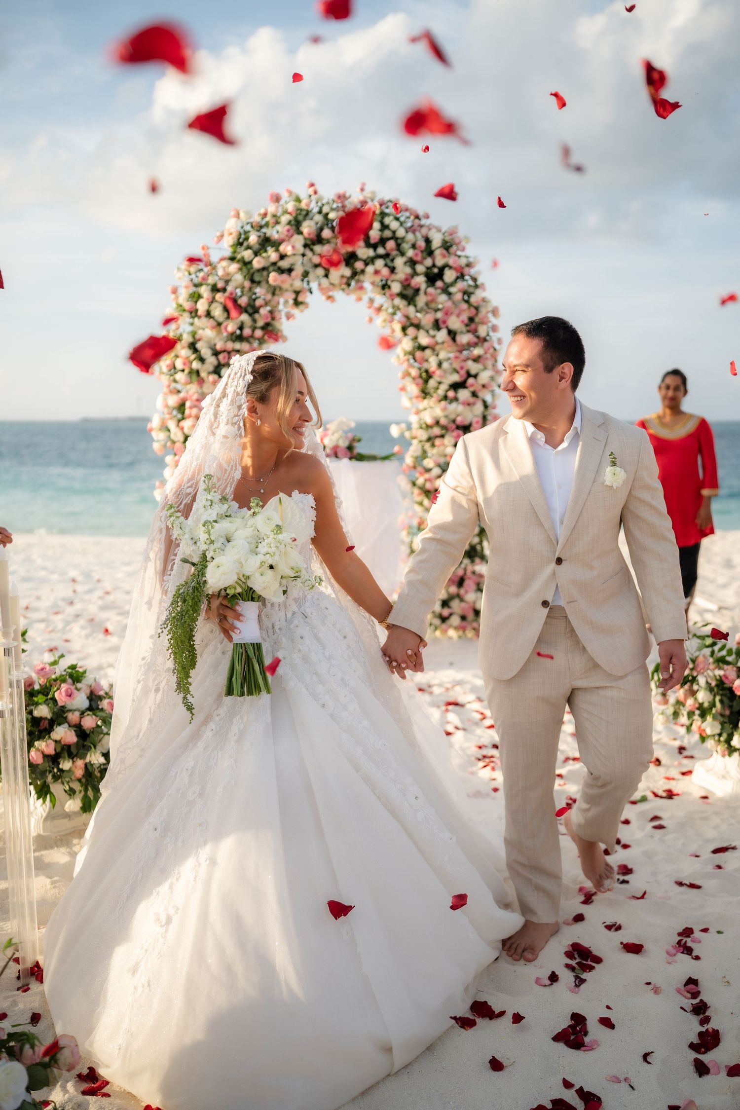 A romantic beach wedding ceremony with a floral arch and red rose petals falling as couple celebrates under cloudy skies.