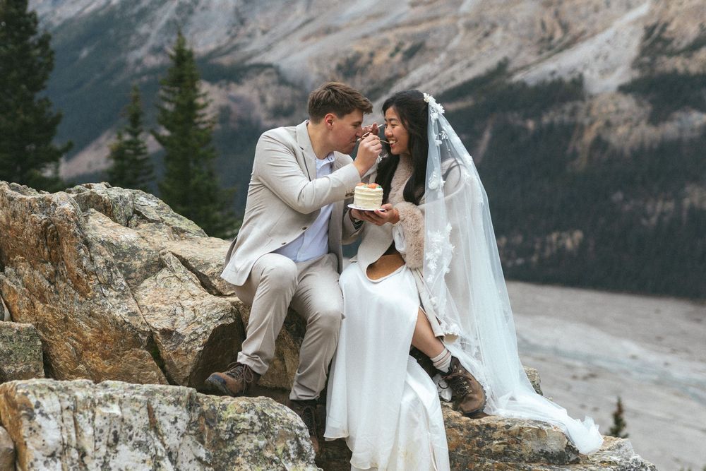 bride and groom laugh and feed each other forkfuls of cake, sitting on a rocky outcrop in the mountains. Both wear hiking boots