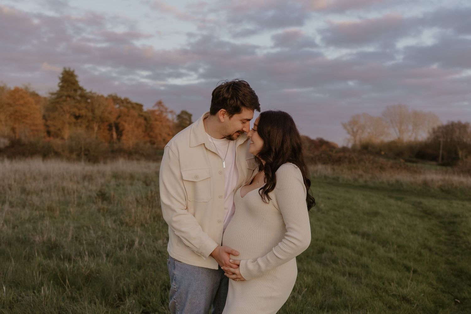 Couple in matching cream outfits embracing during golden hour in a grassy field.