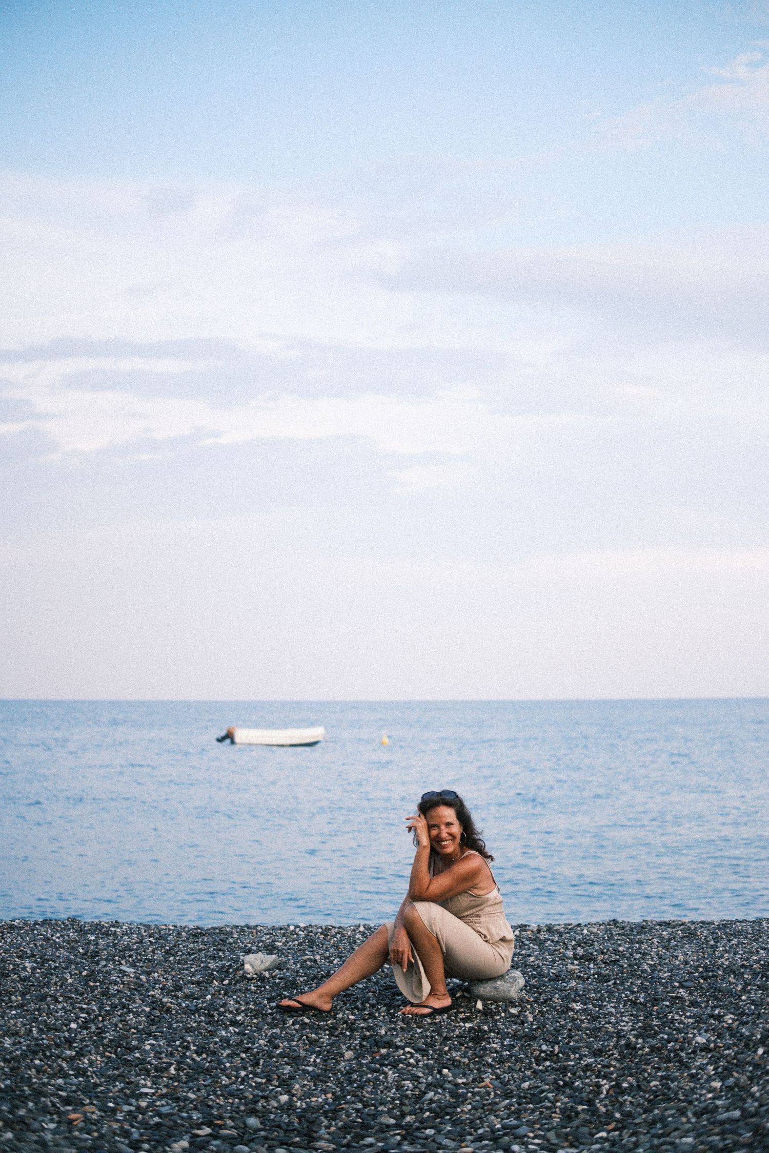 Someone sits on a rocky beach by the calm sea with a small boat visible in the distance on a tranquil day.