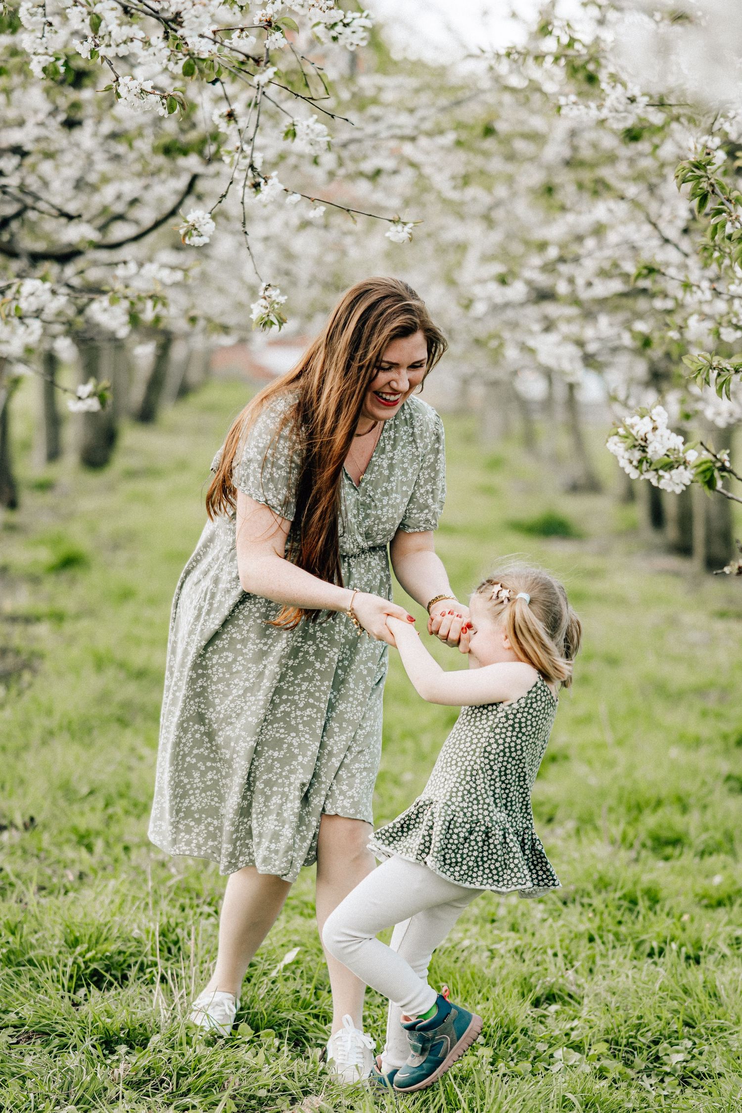 Mother and daughter in matching floral dresses dance together under blooming cherry blossom trees in spring.