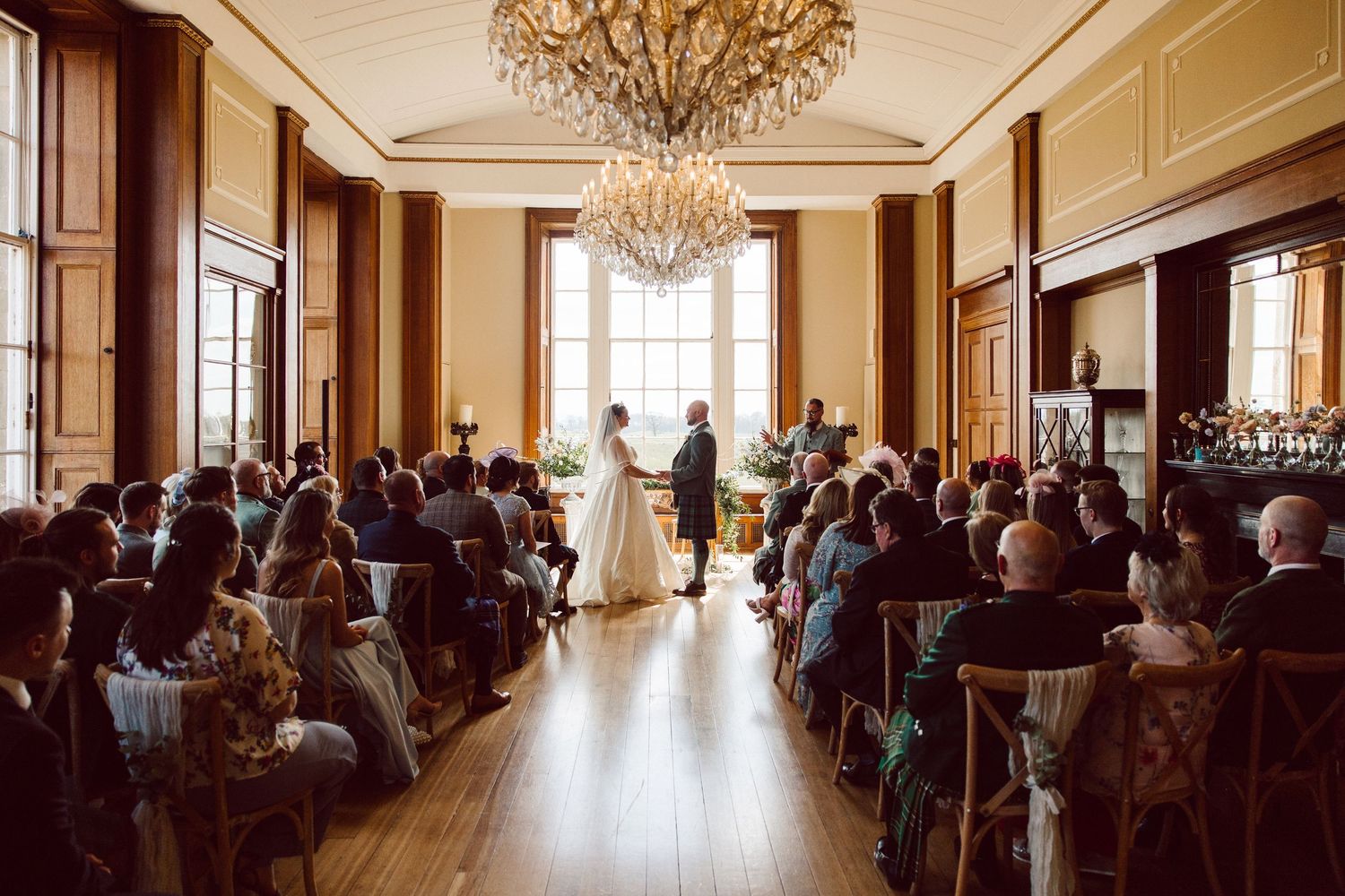 Wedding guests seated in an elegant ballroom with crystal chandeliers and natural light streaming through tall windows.