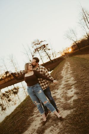 Couple embracing at sunset by a lake on a sandy path, woman smiling with arms outstretched in fall setting.