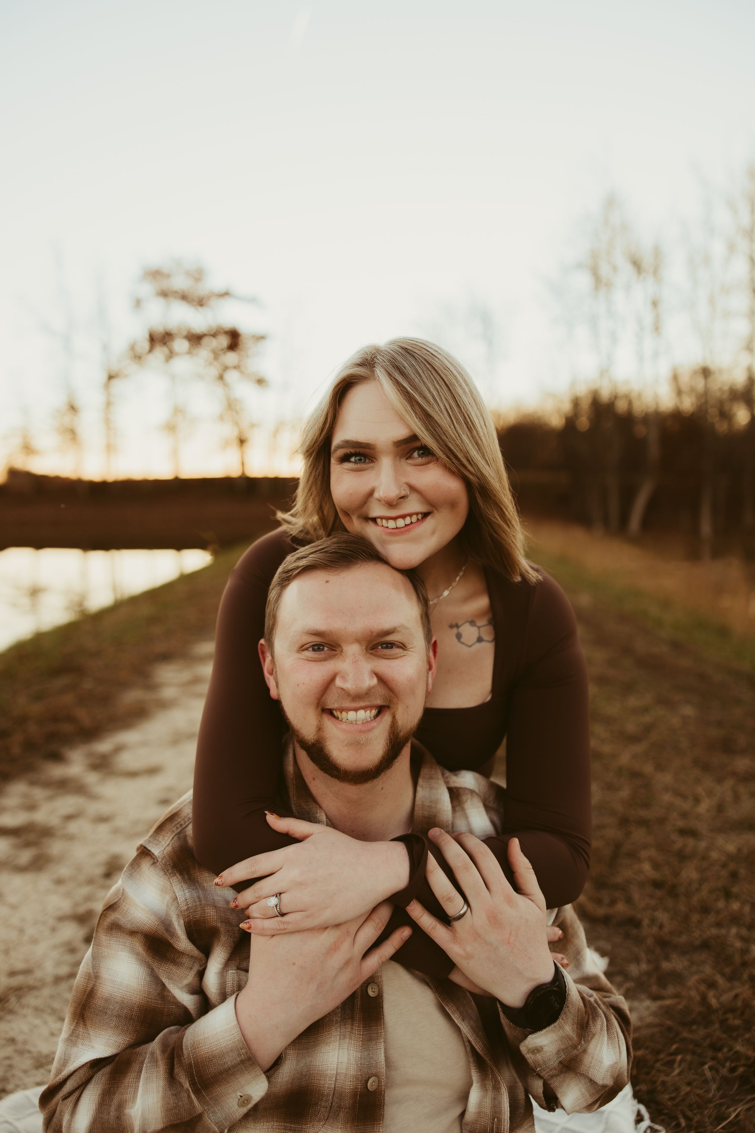 Happy couple posing outdoors at golden hour, woman hugging man from behind showing engagement ring near a lake.