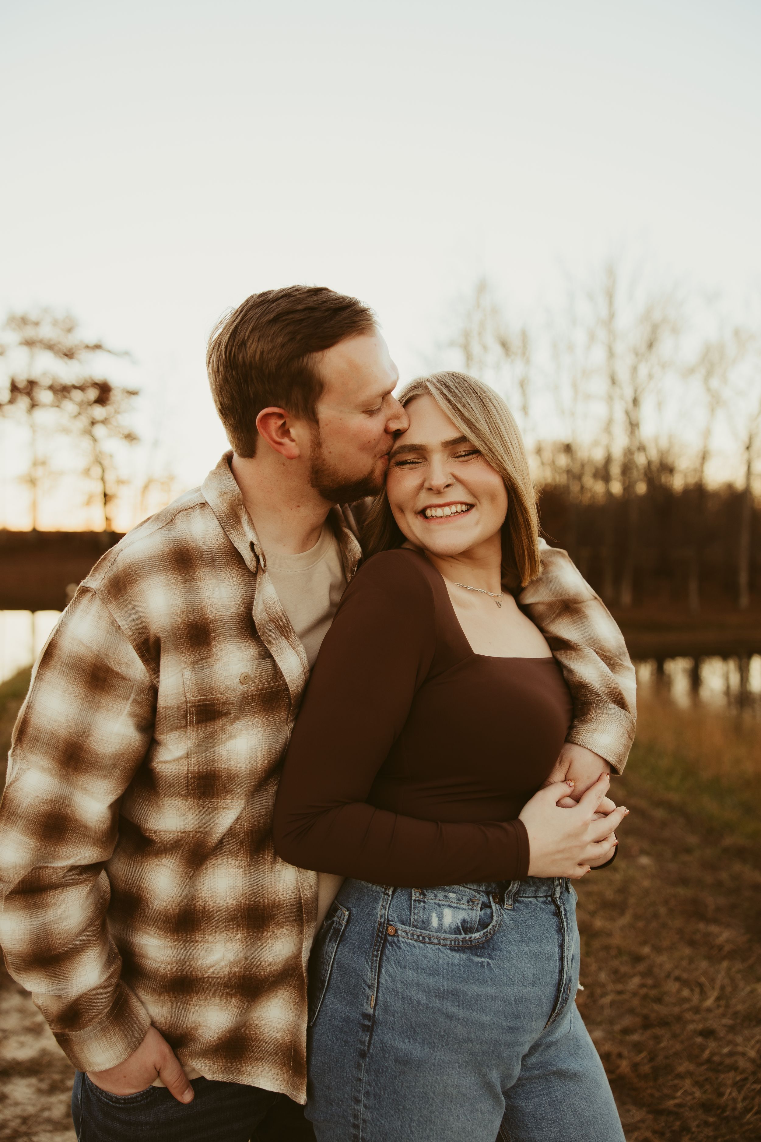 Man kissing woman on cheek at sunset by a lake, woman smiling in brown top and jeans with plaid shirt.