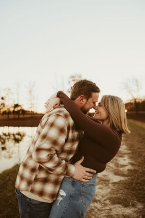 Engaged couple embracing nose-to-nose at golden hour beside a reflective lake surrounded by bare trees.
