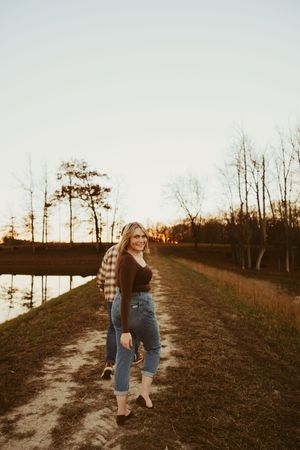Blonde woman walking along a sandy lakeside path at sunset wearing jeans and a brown top with plaid shirt.