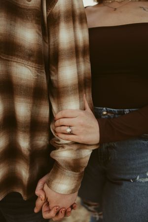 Close-up of couple holding hands showing diamond engagement ring, wearing plaid and brown outfits outdoors.