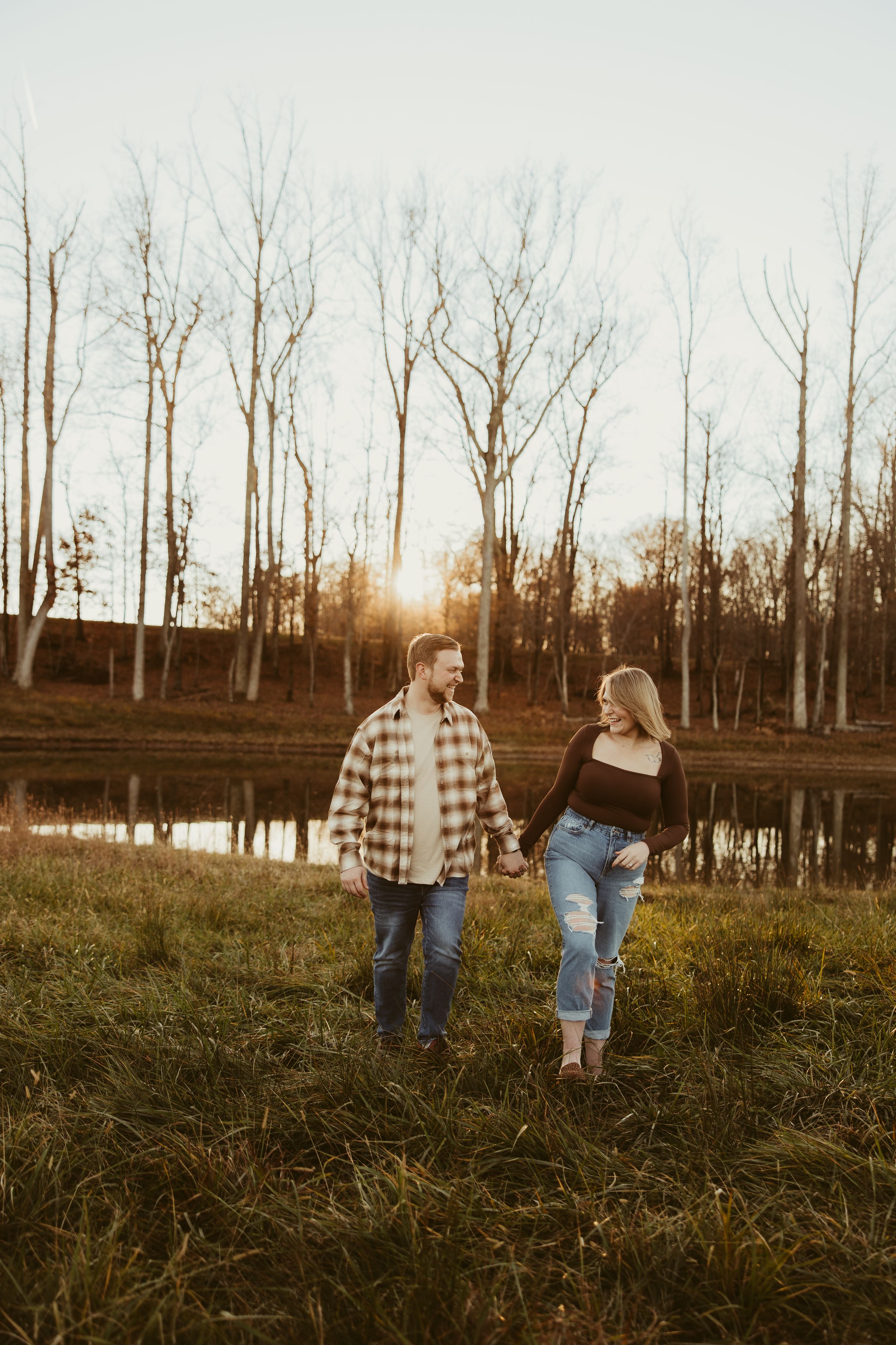 Couple holding hands and walking through a golden field near a pond at sunset with bare trees behind them.