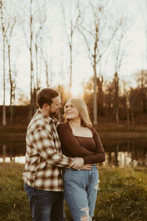 Man embracing woman from behind as she smiles up at him in a sunlit field beside a pond in autumn.