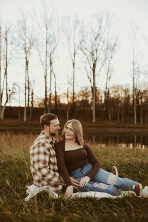 Couple sitting together on a blanket in a golden field at sunset, smiling at each other near a pond.
