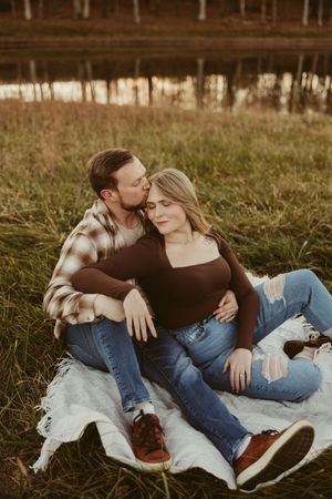 Couple relaxing on a blanket in an autumn field at sunset, man kissing woman on the cheek near a pond.