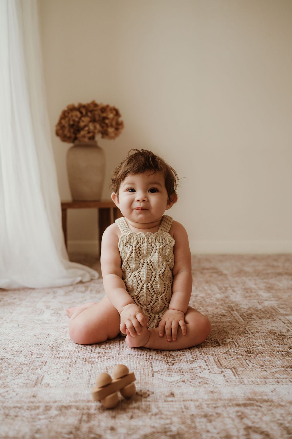 A toddler in a knitted beige romper sits on a textured pink bedspread in a series of candid poses.