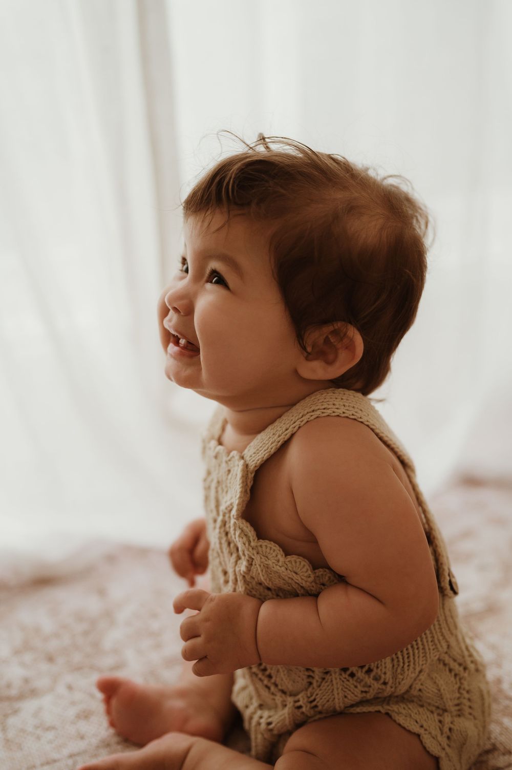 Baby wearing cream knit romper lies on soft carpet while looking up with wonder in dreamy natural light setting.