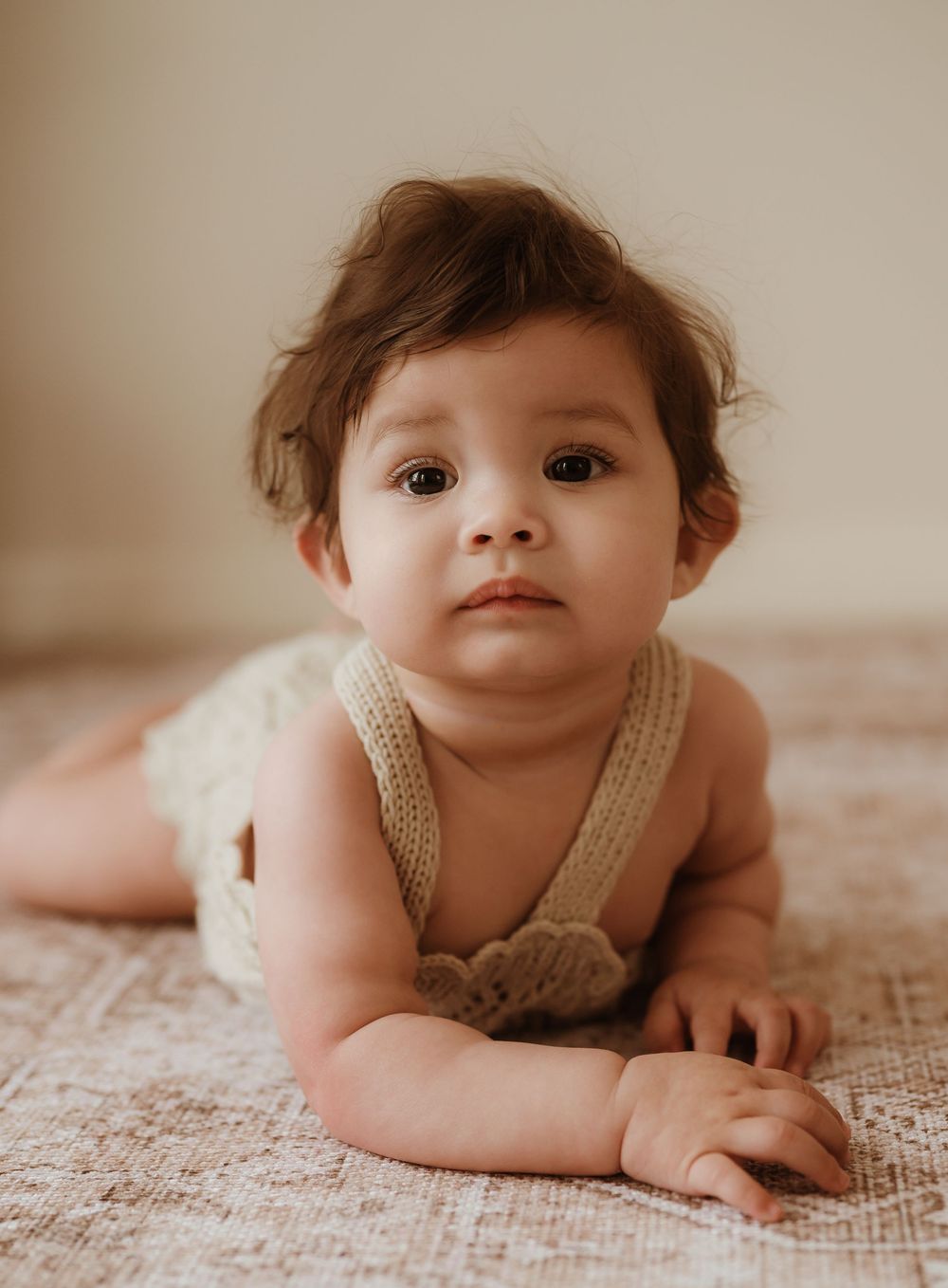A baby wearing a cream-colored knit romper lies on a textured beige blanket in a series of candid poses.