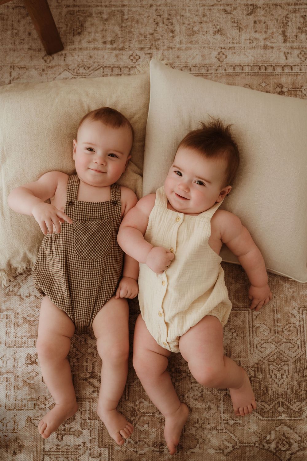 Two babies lying side by side on a beige rug wearing brown and cream colored rompers in a vintage style photo.