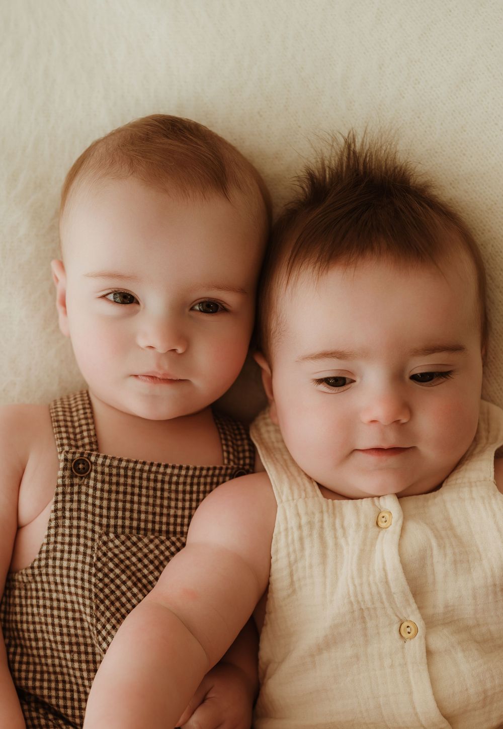 Two babies lying side by side, one wearing a checkered romper and one in a cream colored outfit with buttons.